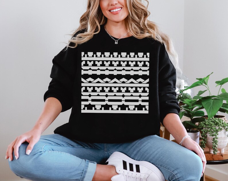 May include: Black sweatshirt with a white geometric pattern across the chest. The pattern consists of various shapes and lines. The model is wearing blue jeans and white sneakers. The sweatshirt is a casual style.
