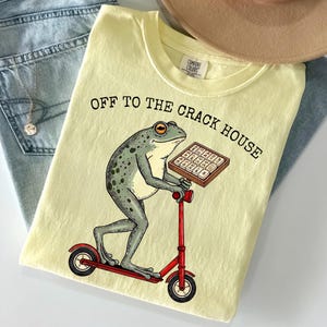 May include: Light yellow t-shirt with the text "OFF TO THE CRACK HOUSE" and a cartoon frog riding a red scooter while holding a box. The shirt is folded on top of blue jeans and a tan hat.