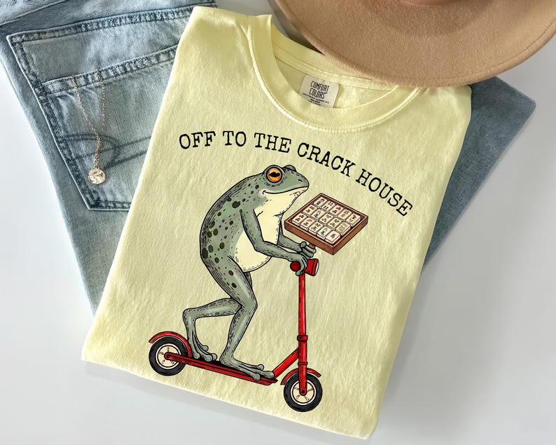 May include: Light yellow t-shirt with the text "OFF TO THE CRACK HOUSE" and a cartoon frog riding a red scooter while holding a box. The shirt is folded on top of blue jeans and a tan hat.