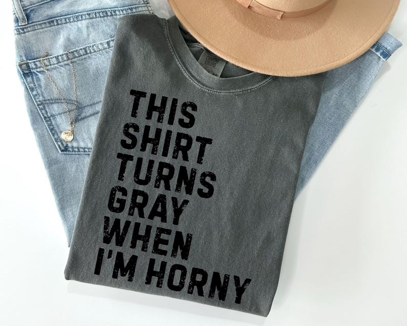 May include: Gray t-shirt with the text "THIS SHIRT TURNS GRAY WHEN I'M HORNY" in black block letters. The shirt is laid on top of light blue jeans and a tan hat. A silver necklace is visible.