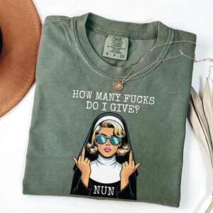 May include: Olive green t-shirt with the text "HOW MANY FUCKS DO I GIVE?" and a graphic of a nun wearing sunglasses and giving the middle finger. The word "NUN" is below the graphic. A gold necklace is draped across the shirt.