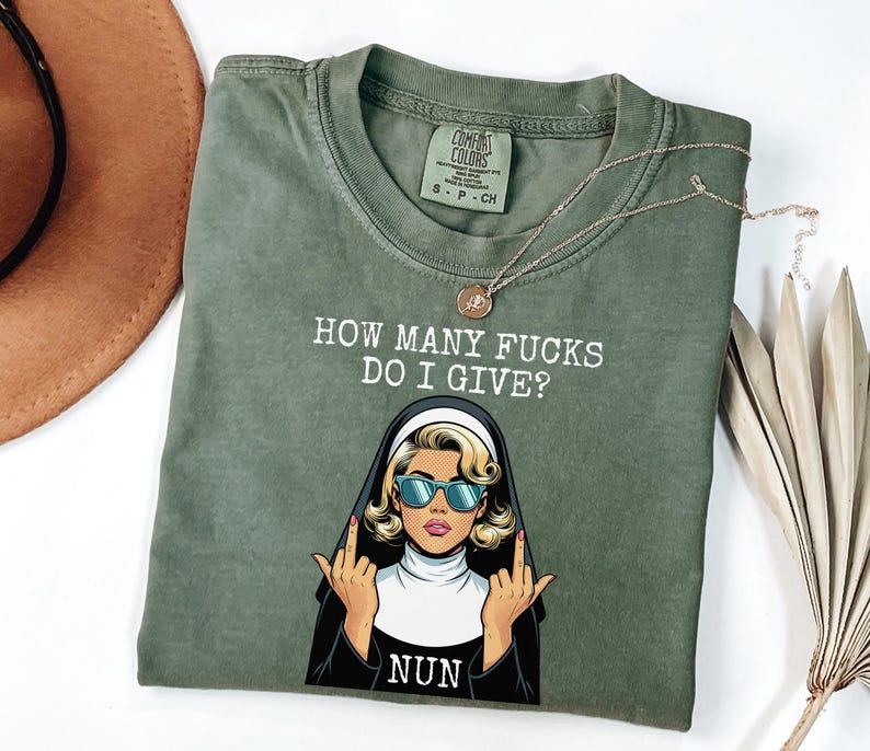 May include: Olive green t-shirt with the text "HOW MANY FUCKS DO I GIVE?" and a graphic of a nun wearing sunglasses and giving the middle finger. The word "NUN" is below the graphic. A gold necklace is draped across the shirt.