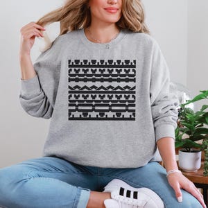 May include: A light gray sweatshirt with a black geometric pattern. The pattern is composed of various shapes, including rectangles, triangles, and lines. The sweatshirt is a crew neck style.