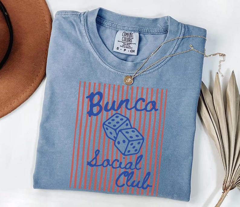 May include: A blue short-sleeve t-shirt with a Comfort Colors label at the neck. The front features a graphic with vertical orange stripes, two dice, and the blue text Bunco Social Club. A gold-toned necklace rests on the collar of the shirt.