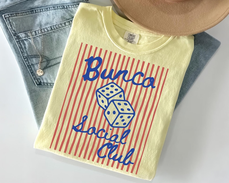 May include: A pale yellow t-shirt featuring a graphic print of two dice and the blue text "Bunco Social Club" centered over a background of vertical red stripes. The shirt has a crew neckline and a Comfort Colors brand label at the inner collar.