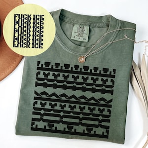 May include: An olive green t-shirt with a black geometric design and the text "FUCK ICE" in a vertical pattern. A gold necklace with a pendant is draped on the shirt. The shirt is a Comfort Colors brand.