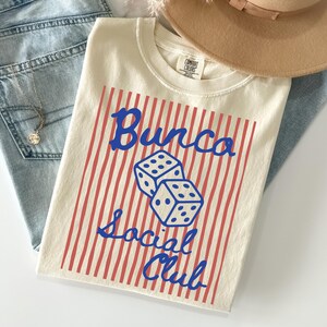 May include: A cream-colored t-shirt featuring a red vertical striped background with two blue dice in the center. The blue text reads Bunco Social Club. The shirt has a crew neckline and a Comfort Colors brand label on the inside of the collar.