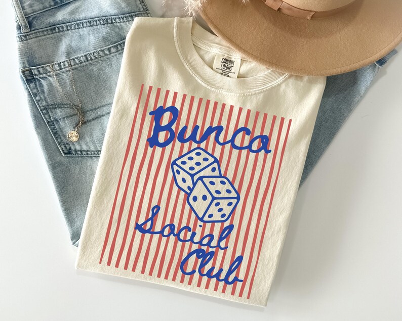 May include: A cream-colored t-shirt featuring a red vertical striped background with two blue dice in the center. The blue text reads Bunco Social Club. The shirt has a crew neckline and a Comfort Colors brand label on the inside of the collar.