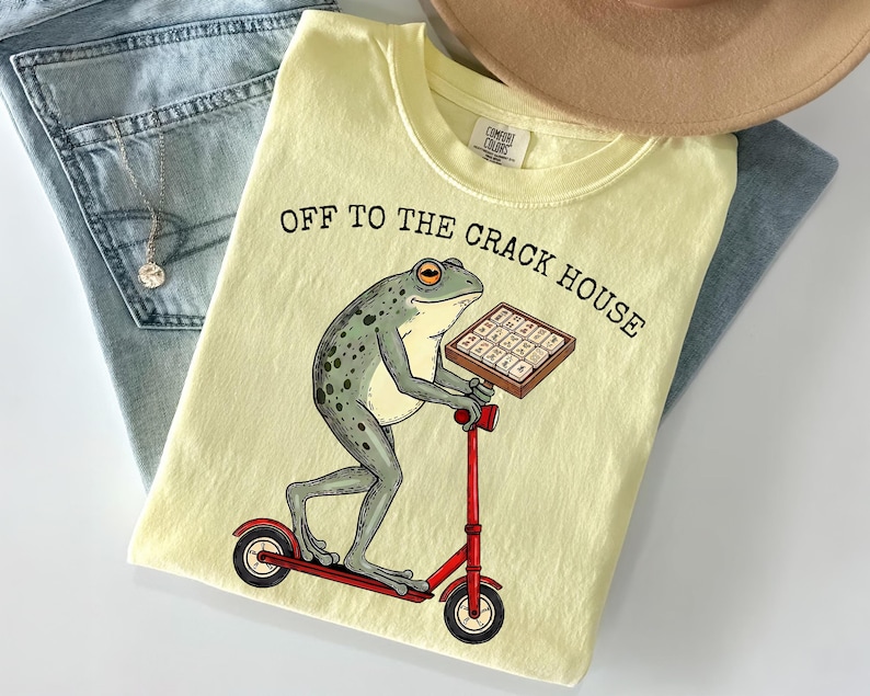 May include: Light yellow t-shirt with the text "OFF TO THE CRACK HOUSE" and a cartoon frog riding a red scooter while holding a box. The shirt is folded on top of blue jeans and a tan hat.