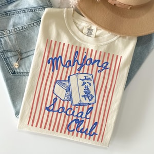 May include: Cream-colored t-shirt with a blue and red design. The shirt features the words "Mahjong Social Club" and a graphic of two Mahjong tiles. The shirt is paired with light blue jeans and a tan hat.