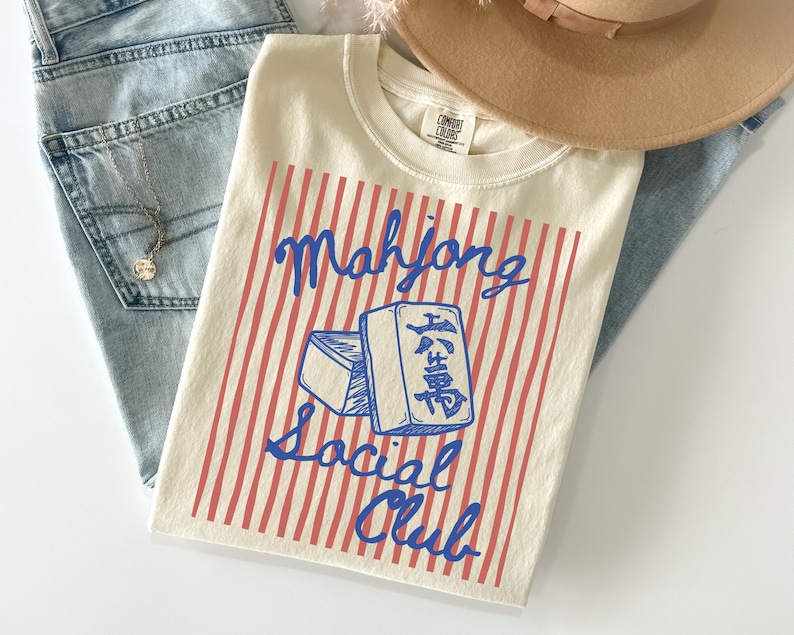 May include: Cream-colored t-shirt with a blue and red design. The shirt features the words "Mahjong Social Club" and a graphic of two Mahjong tiles. The shirt is paired with light blue jeans and a tan hat.