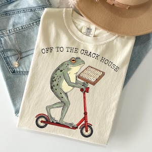May include: Cream-colored t-shirt featuring a frog riding a red scooter and holding a tray of what appears to be brownies. The text "OFF TO THE CRACK HOUSE" is printed above the frog. A tan hat and blue jeans are in the background.