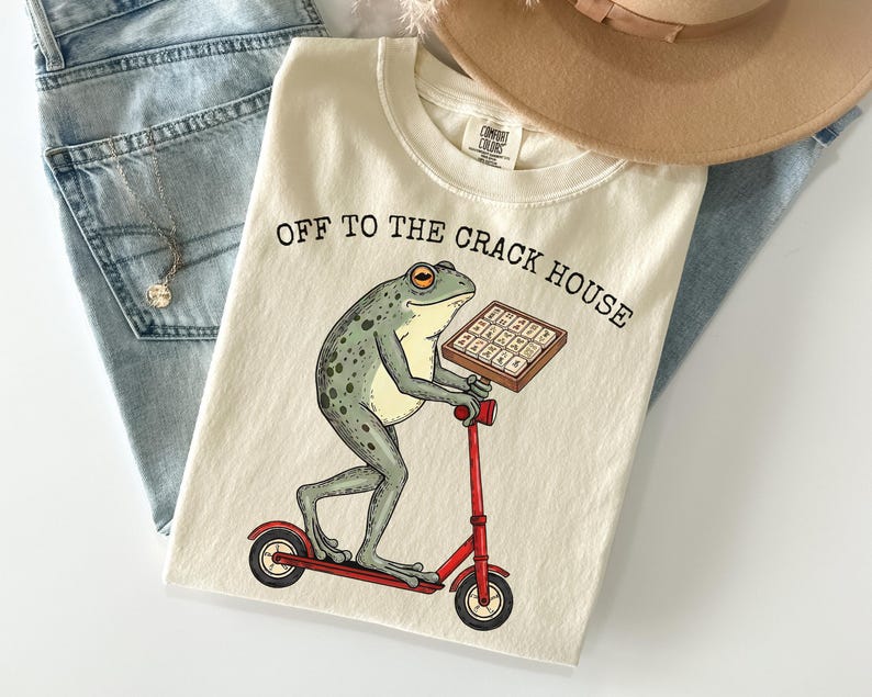 May include: Cream-colored t-shirt featuring a frog riding a red scooter and holding a tray of what appears to be brownies. The text "OFF TO THE CRACK HOUSE" is printed above the frog. A tan hat and blue jeans are in the background.