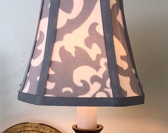 May include: A gray and white patterned lampshade with a gray trim. The lampshade is a drum shape and is lit by a light bulb.