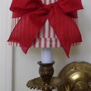 Puede incluir: Una pantalla de lámpara a rayas rojas y blancas con un lazo rojo. La pantalla está unida a una pared de metal dorado.