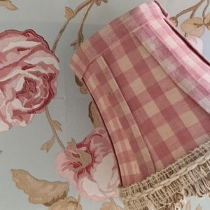 May include: A pink and white gingham lampshade with a ruffled trim. The lampshade is made of fabric and has a soft, feminine look.