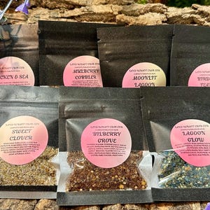 May include: Seven black resealable pouches with clear windows containing various granular mixtures. Each pouch features a round pink label with the text Land Scraft Crab Co and specific scent names including Chicken & Sea, Mulberry Cobbler, Moonlit Lagoon, Birds of a Feather, Sweet Clover, Bilberry Grove, and Lagoon Glow.
