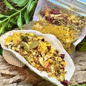 May include: A white seashell filled with a mixture of yellow powder, green pumpkin seeds, and dried red flower petals. A clear, iridescent plastic pouch containing the same mixture of yellow powder, seeds, and dried petals sits behind the shell.