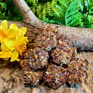 May include: A pile of brown, round treats, speckled with colorful dried flowers and herbs. The treats are arranged on a textured, brown surface, with a yellow flower and a piece of wood in the background.