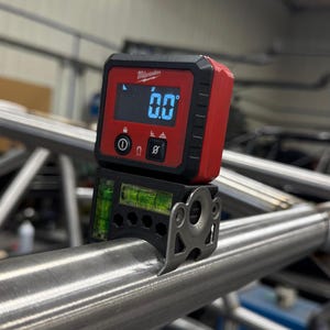 May include: A red and black digital level by Milwaukee displays 0.0 degrees on its screen. The level is mounted on a silver metal tube, with a bubble level visible below the digital display. The tool is designed for precision measurement.