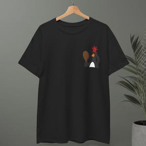 Puede incluir: Camiseta negra con un diseño de pingüino de dibujos animados. El pingüino sostiene una paleta de ping-pong y una corona roja. La camiseta está colgada en una percha de madera.