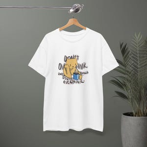 Puede incluir: Camiseta blanca con un gato de dibujos animados sosteniendo una taza de café azul y el texto "DON'T OVERTHINK". El gato es de color marrón claro con una expresión neutra. La camiseta está colgada en una percha de madera.