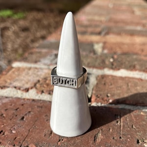May include: A silver ring with the word "BUTCH" engraved on it. The ring is displayed on a white ring holder.