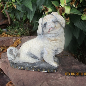 May include: A white concrete garden statue of a dog sitting on a rock. The dog has a black nose and eyes, and a black stripe down its back.
