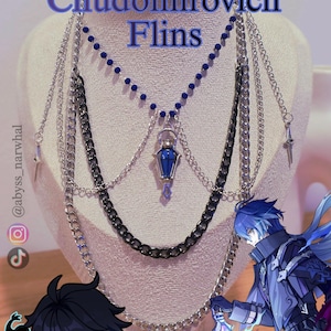 May include: A multi-layered necklace display featuring silver and black chains, a blue beaded strand, and a blue pendant. The text "Kyryll Chudomirovich Flins" is displayed at the top. The display is on a mannequin head.