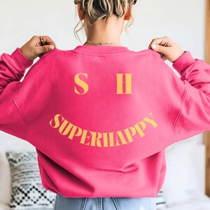 May include: A bright pink sweatshirt with the text "SUPERHAPPY" in yellow, forming a smiling face. The sweatshirt also features the letters "S" and "H" above the smile. The model is wearing blue jean shorts.