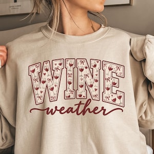 Könnte beinhalten: Ein sandfarbenes Sweatshirt mit Rundhalsausschnitt und dem Wort WINE in großen Blockbuchstaben, die mit einem Muster aus Weingläsern, Herzen und Schleifen gefüllt sind. Darunter steht das Wort weather in dunkelroter Schreibschrift.