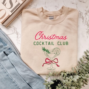 May include: A beige sweatshirt with the text "Christmas Cocktail Club" in red and green. A cocktail glass graphic with a red bow is below the text. The sweatshirt is displayed on a marble surface with denim jeans and greenery.