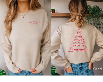 May include: Sand-colored sweatshirt with long sleeves. The front features the text "Merry Christmas" in pink script. The back displays a pink Christmas tree design with a star on top. The model is wearing blue jeans.