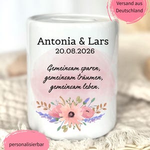 以下が含まれることがあります： Antonia & Lars 20.08.2026というテキストとドイツ語のフレーズGemeinsam sparen, gemeinsam tr&auml;umen, gemeinsam lebenが印刷された白い陶器製の貯金箱。淡いピンクの水彩画風の背景と花のイラストが描かれています。