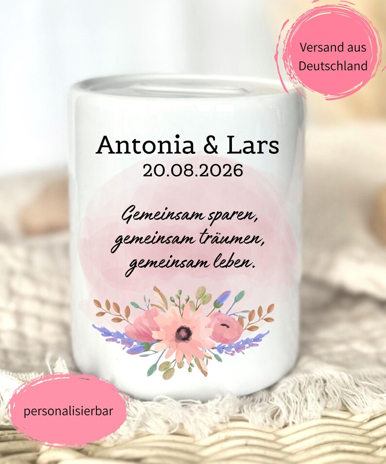 以下が含まれることがあります： Antonia & Lars 20.08.2026というテキストとドイツ語のフレーズGemeinsam sparen, gemeinsam tr&auml;umen, gemeinsam lebenが印刷された白い陶器製の貯金箱。淡いピンクの水彩画風の背景と花のイラストが描かれています。
