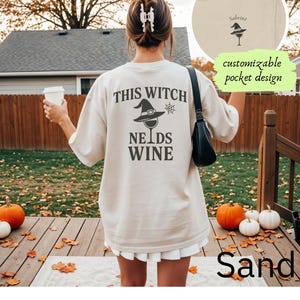 May include: Sand-colored t-shirt with the text "THIS WITCH NEEDS WINE" in black. The design includes a witch hat and a wine glass. The shirt has a customizable pocket design and is perfect for Halloween.