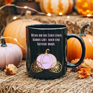 Könnte beinhalten: Schwarze Keramik-Tasse mit Henkel, mit dem Text "Wenn dir das Leben einen Kürbis gibt. Mach eine Kutsche draus." und einem rosa Kürbis-Kutschen-Design mit goldenen Rädern. Der Hintergrund zeigt Kürbisse und Heu.