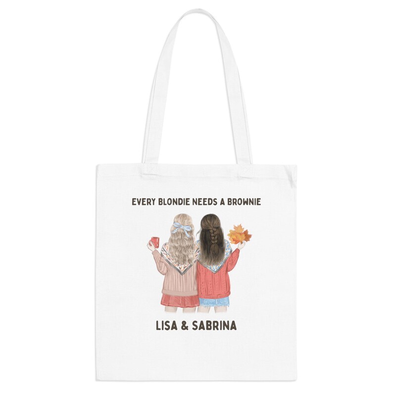 May include: White tote bag featuring an illustration of two women with the text "EVERY BLONDIE NEEDS A BROWNIE" and the names "LISA & SABRINA". One woman holds a mug, the other holds fall leaves. The women are wearing sweaters and shorts.
