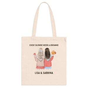 May include: Cream-colored tote bag featuring an illustration of two women with the text "EVERY BLONDIE NEEDS A BROWNIE" and the names "LISA & SABRINA". One woman holds a mug, the other holds fall leaves.
