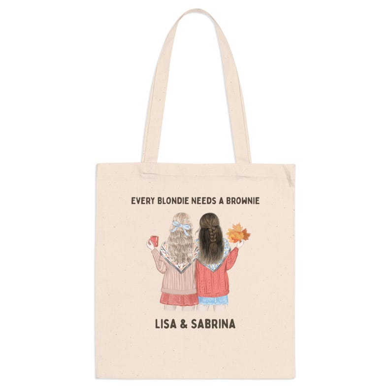 May include: Cream-colored tote bag featuring an illustration of two women with the text "EVERY BLONDIE NEEDS A BROWNIE" and the names "LISA & SABRINA". One woman holds a mug, the other holds fall leaves.