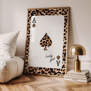 May include: A framed wall art print featuring the Ace of Spades playing card design. The card's spade symbol and border have a leopard print pattern. The text "Lucky girl" is written on the card. The frame is gold.