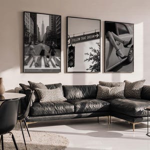 Op de afbeelding: Drie ingelijste zwart-witfoto's hangen boven een zwarte leren bank. De foto's tonen stadsgezichten en een auto met benen uit het raam. De middelste foto heeft de tekst "FOLLOW THAT DREAM". Kussens met luipaardprint.