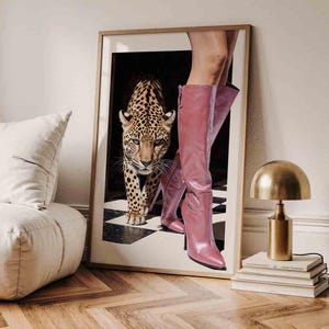 May include: A framed art print showing a leopard walking towards the viewer, with pink knee-high boots in the foreground. The leopard has a spotted coat. The boots are a vibrant pink colour and have a pointed toe.