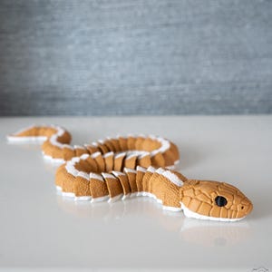 May include: A tan-coloured snake toy with white detailing along its back. The snake has a segmented body and a detailed head with black eyes. The toy is designed to resemble a real snake and is placed on a white surface.