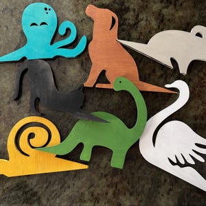May include: A collection of wooden animal cutouts in various colors. Includes a teal octopus, brown dog, gray mouse, black cat, yellow snail, green dinosaur, and white swan. These decorative pieces are likely for crafts or home decor.