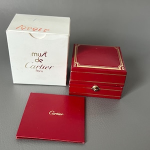 Könnte beinhalten: Eine weiße Schachtel mit dem Text "must de Cartier Paris" und eine rote Schachtel mit goldenen Verzierungen sowie eine kleine rote Karte mit dem Cartier-Logo. Die Artikel befinden sich auf einer grauen Oberfläche.