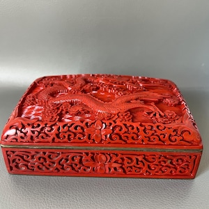 May include: A vibrant red, rectangular box with intricate carvings. The top features a detailed dragon design, while the sides showcase floral and scroll patterns. The box has a brass-colored trim.