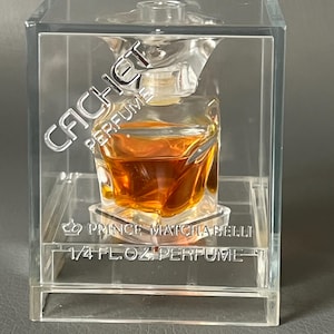Può includere: Un piccolo flacone di profumo trasparente con liquido ambrato, racchiuso in una scatola di plastica trasparente. Il testo "CACHET PERFUME" è visibile sulla scatola, insieme a "PRINCE MATCHABELLI" e "1/4 FL.OZ. PERFUME".