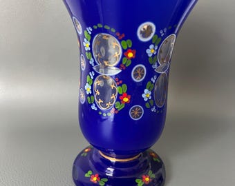 Op de afbeelding: Een kobaltblauwe glazen vaas met een gouden rand en voet. De vaas is versierd met heldere, cirkelvormige ontwerpen en kleine bloemmotieven in rood, geel en blauw. De vaas heeft een uitlopende bovenkant en een afgeronde voet.