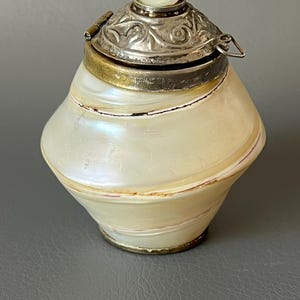 Pode incluir: Um recipiente perolado em forma de concha com uma tampa de prata articulada e ornamentada. A tampa apresenta um pequeno detalhe oval de madrepérola. O recipiente tem uma faixa de latão e é provavelmente um item decorativo.